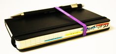 a black notebook with a purple ribbon tied around the cover and two pens on top