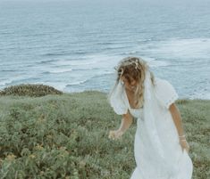 a woman in a white dress is walking by the ocean with her hair blowing in the wind