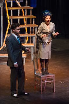 a woman standing on top of a wooden chair next to a man in a suit