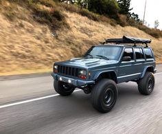 a blue jeep driving down the road with a surfboard on top of it's roof