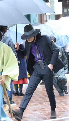 a man in a suit and hat is holding an umbrella on the sidewalk while people walk by