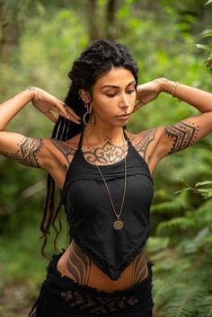 a woman with tattoos on her arms and chest standing in front of some trees wearing a black top