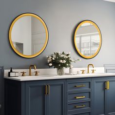 two round mirrors are above the double sinks in this gray and white bathroom with gold accents