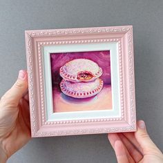 a painting of two pies in a pink frame on a gray wall with hands holding it