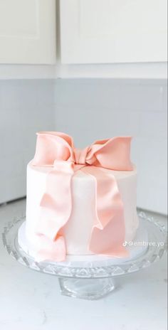 a white and pink cake with a bow on the top is sitting on a glass plate