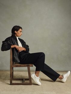 a man sitting in a chair with his legs crossed, wearing white sneakers and a black jacket