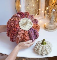 a hand holding a crocheted hat next to a white pumpkin on a table