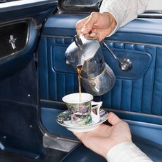 a person pours coffee from a teapot into a cup in front of a blue car