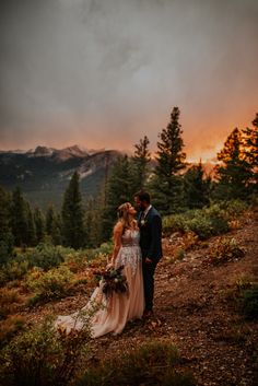 a bride and groom standing on top of a mountain at sunset