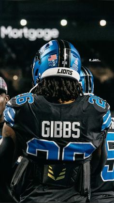 two football players sitting on the sidelines during a game, one is wearing a black and blue uniform with lions written on it