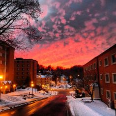 the sky is pink and orange as it sets on a snowy street in front of some buildings