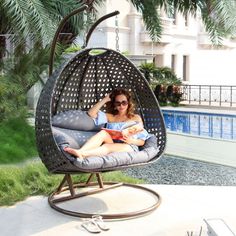 a woman sitting in a hanging egg chair next to a swimming pool and holding a book