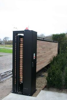 a large black box sitting on top of a sidewalk next to a fence and bushes