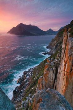 the sun is setting over an ocean with rocks and mountains in the foreground,