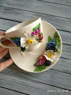 a hand holding a white cup and saucer with colorful flowers on the side, sitting on a wooden table