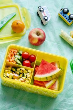 a lunch box filled with fruits and vegetables on top of a blue table cloth next to plastic utensils