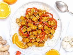 a white plate topped with orange chicken and rice next to sliced oranges, garlic and ginger