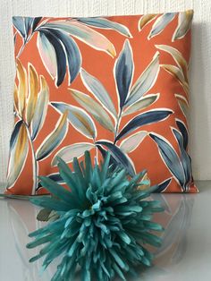 an orange pillow with blue flowers on it next to a green flower in front of a white wall