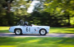 an old race car is driving down the road in front of some green grass and trees