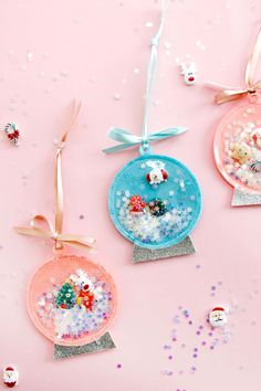 three snow globe ornaments hanging from ribbons on a pink background with other decorations around them