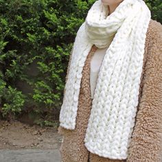 a woman wearing a white and brown knitted scarf in front of some bushes with green leaves