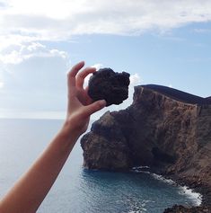 a person holding up a rock in front of an ocean and cliff side with blue sky