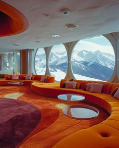 an orange couch in front of large round windows with snow covered mountains behind it,