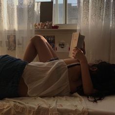 a woman laying on her side reading a book in front of a window with curtains