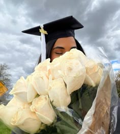 a woman wearing a graduation cap and gown holding flowers in front of her face with cloudy skies behind her