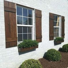 three windows with wooden shutters on the side of a white brick building and green bushes in front