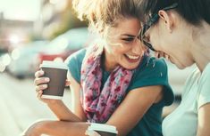 two women are laughing while looking at a cell phone and holding a cup of coffee