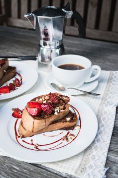 two white plates topped with french toast covered in strawberries next to a cup of coffee