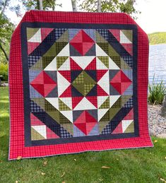 a red and black quilt is on the grass near some water with trees in the background