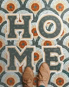 someone's feet in brown shoes standing on a colorful tile floor with the word home