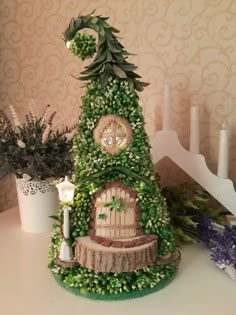 a small house made out of flowers and greenery sits on a table next to candles