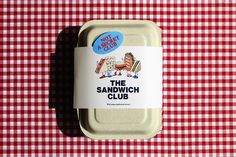 the sandwich club lunch box sits on a red and white checkered tablecloth with an advertisement for sandwiches