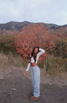 a woman standing in the dirt with her hands behind her head and looking at the camera