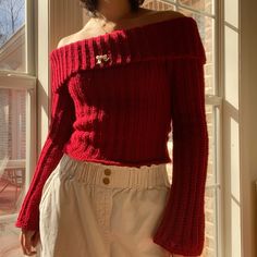 a woman standing in front of a window wearing a red off the shoulder sweater and white shorts