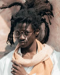 a woman with glasses and dreadlocks standing in front of a stone wall wearing an orange scarf