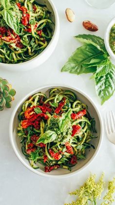 two white bowls filled with pasta and vegetables next to some green leaves on the table