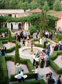 a group of people standing in front of a fountain surrounded by hedges and bushes, all looking at each other