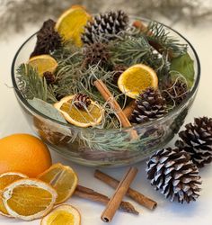 oranges, pine cones and cinnamon sticks are arranged in a glass bowl on a white surface