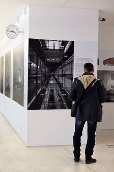 a man standing in front of a wall with pictures on it and a clock hanging from the ceiling