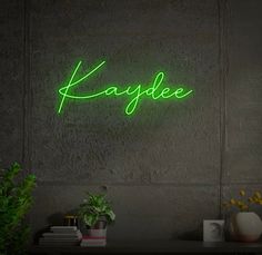 a green neon sign that reads kaydee on the side of a wall next to a potted plant