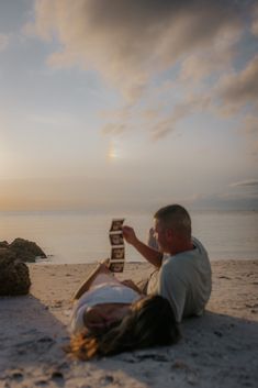 a man and woman laying on the beach with a book in their hands, reading