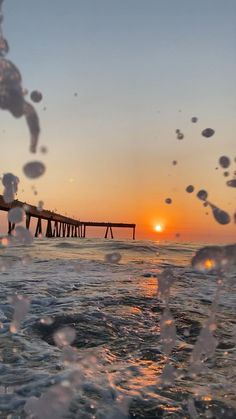 the sun is setting over an ocean with water splashing on it and a pier in the distance