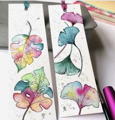 two bookmarks with watercolor flowers on them next to a purple pen and pencil