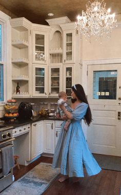 a woman in a blue dress holding a baby while standing in a kitchen with white cabinets