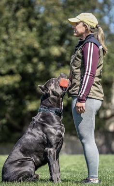 a woman standing next to a black dog on top of a lush green field