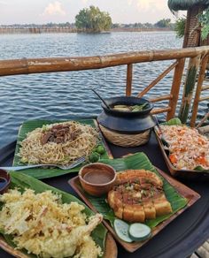 an assortment of food sitting on top of a table next to the water in front of a wooden fence